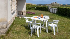 A round table with white chairs set outdoors, surrounded by a grassy area with flowers and a view of the sea and hills in the background. - Ar Wellen by Interhome (Cléder)