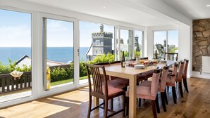Een ruime eethoek met een grote tafel en stoelen, grote ramen met uitzicht op de zee en een kasteelachtig gebouw.
