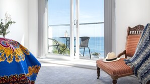 Een kamer met een grote glazen schuifdeur die uitkomt op een balkon met uitzicht op de oceaan. Er staat een rieten stoel met een hoed erop en een kleurrijke deken op de vloer.