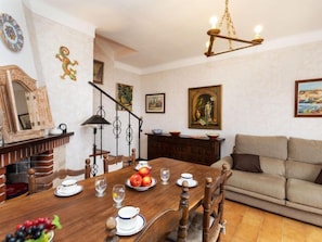 Un comedor con una mesa de madera dispuesta para cuatro personas, rodeada de sillas. Hay una chimenea con repisa de ladrillo, un candelabro y obras de arte en las paredes.