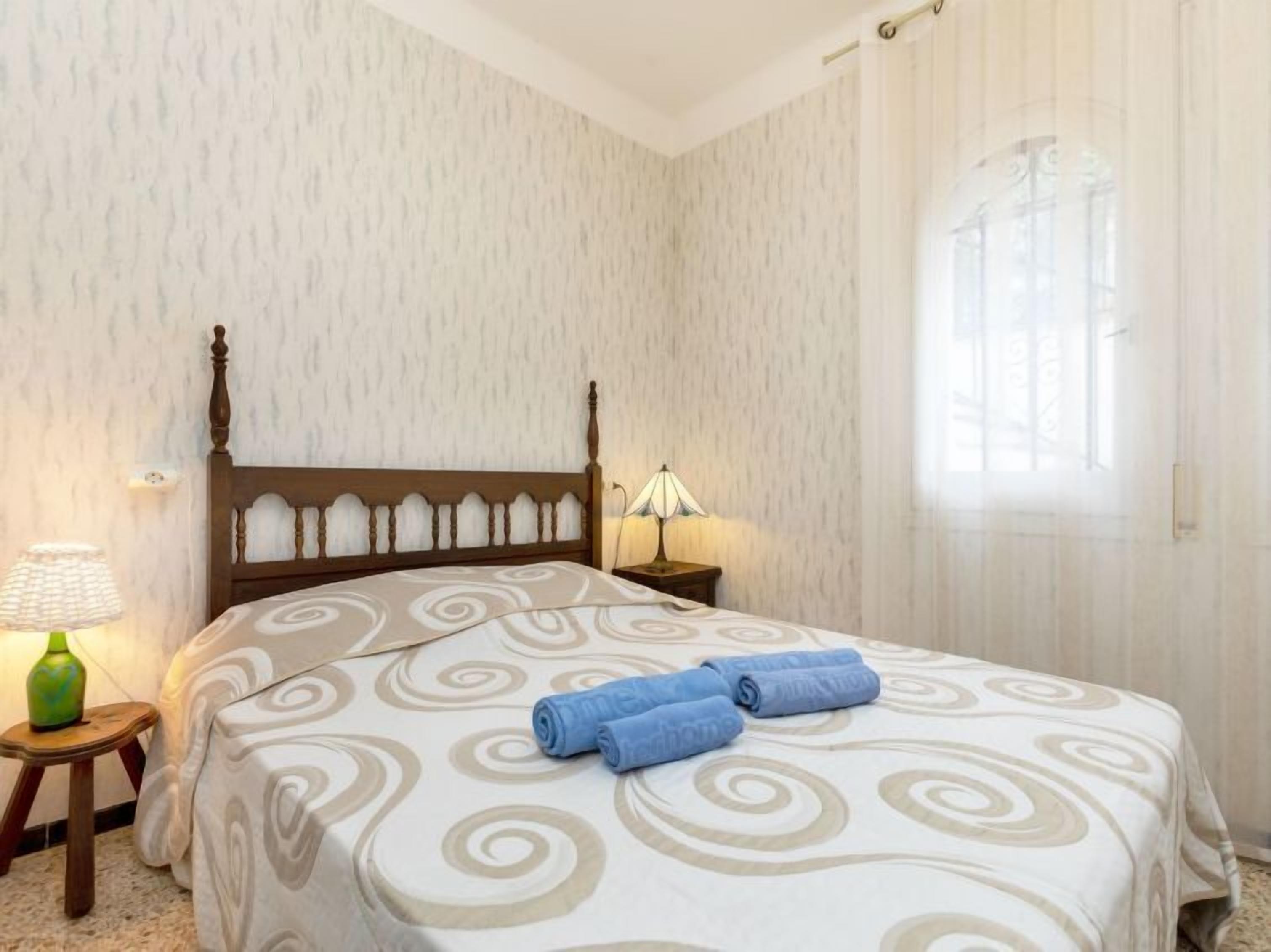 Een slaapkamer met een houten hoofdbord, een bed met een beddekleed met dessin, een bedlampje en een raam met gordijnen.