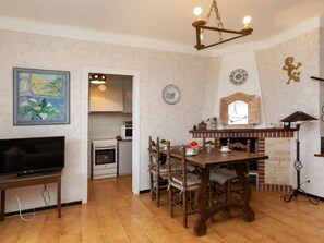 Een eetgedeelte met een houten tafel en stoelen, een televisie op een meubel, een keuken op de achtergrond, en een decoratieve wand met een klok en borden.