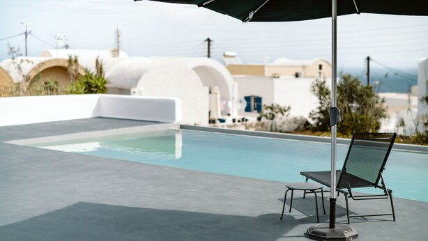 Zona de piscina amb una cadira relaxant i una ombra, una piscina nítida i edificis blancs amb arcs al fons.