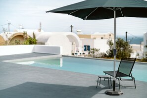 Zona junto a la piscina con una silla de descanso y sombrilla, una piscina cristalina y edificios blancos con arcos al fondo.