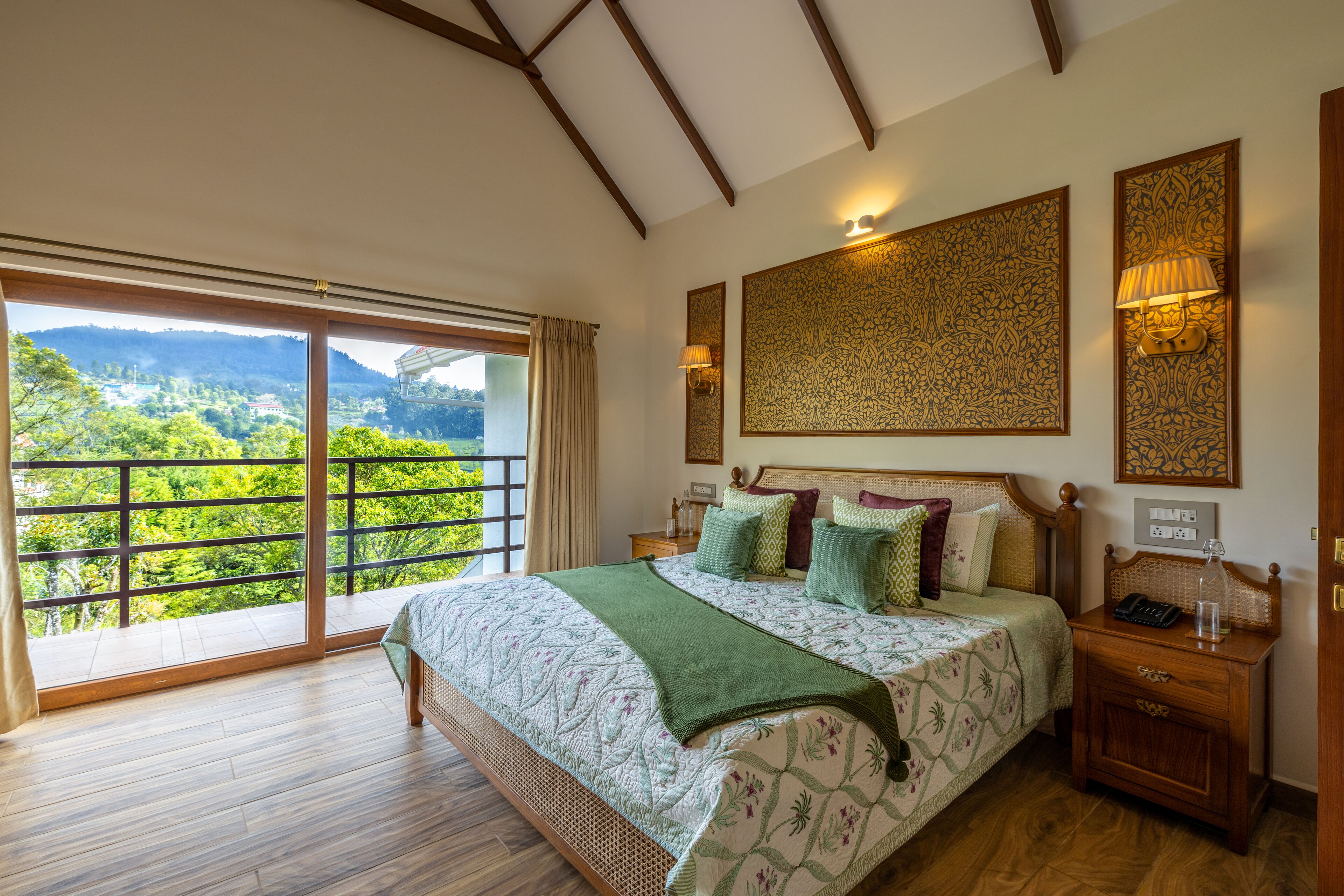 A spacious bedroom with a large bed, a nightstand, a wall-mounted lamp, and a view of lush greenery through a sliding glass door.