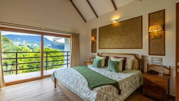 A spacious bedroom with a large bed, a nightstand, a wall-mounted lamp, and a view of lush greenery through a sliding glass door.