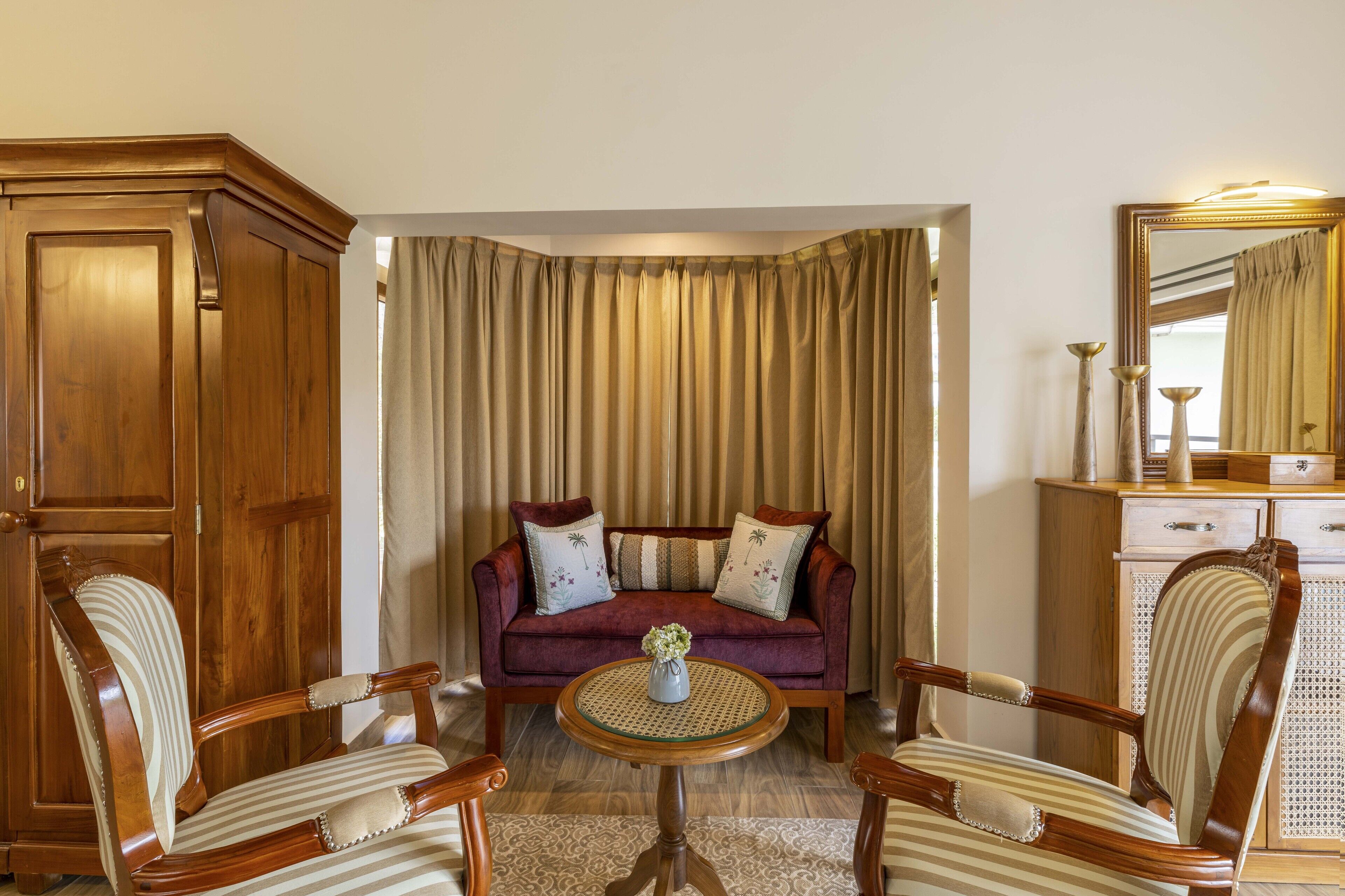 A room with a wooden cabinet, a red sofa with patterned cushions, two striped armchairs, and a small round table with a vase.