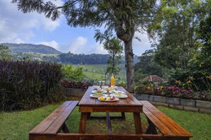 A wooden picnic table set outdoors with plates of food and a bottle of orange juice. - Amã Stays & Trails Werifesteria, Coonoor (Coonoor)