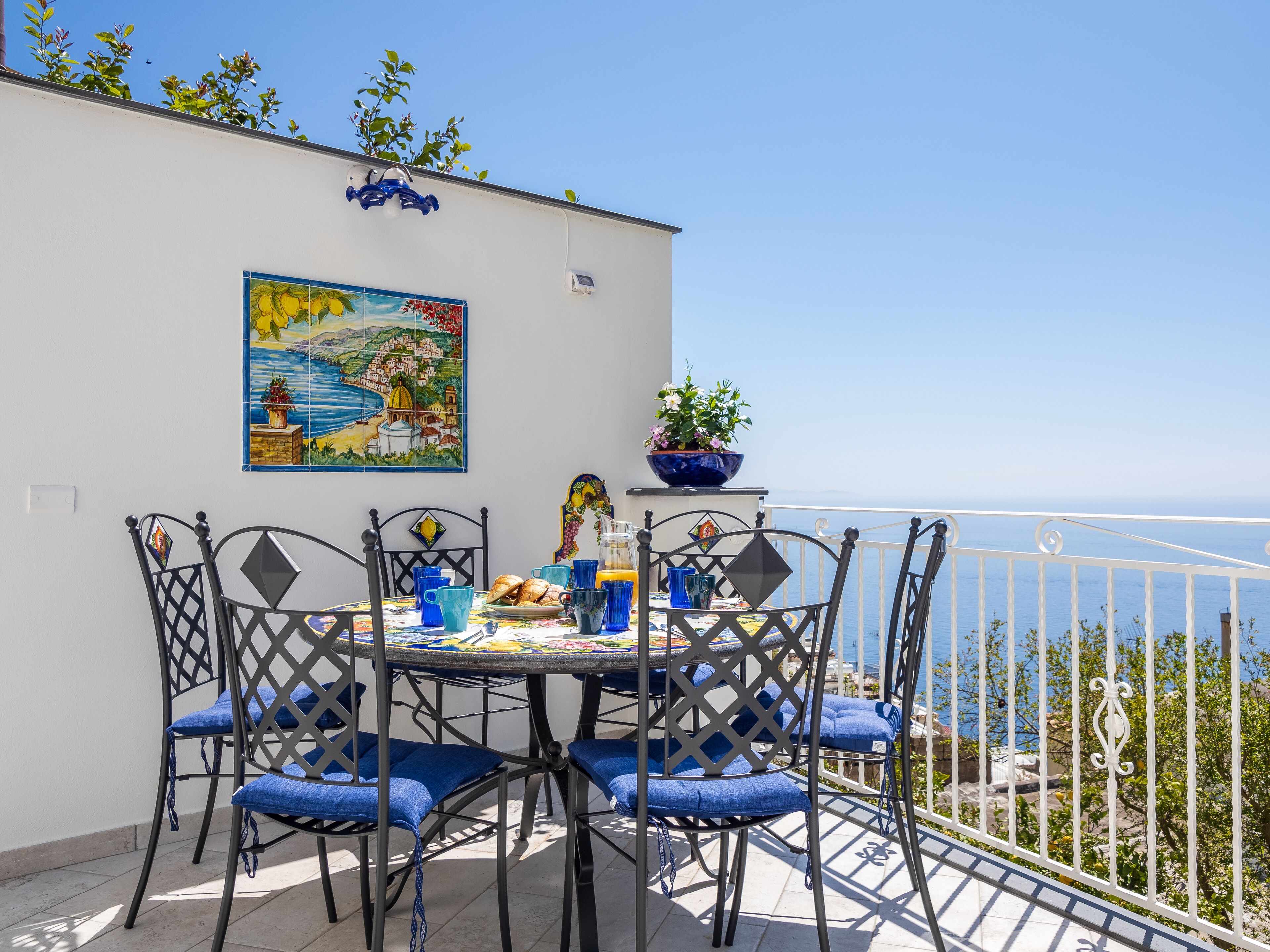 Una terrazza con un tavolo apparecchiato per due, caratterizzata da una tovaglia a scacchi blu e bianca, sedie imbottite blu e un vaso con una pianta.