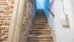 A narrow staircase with a brick wall on one side and a white wall on the other, leading to a room with a visible light fixture. - Rialto al Manganer (Venice)
