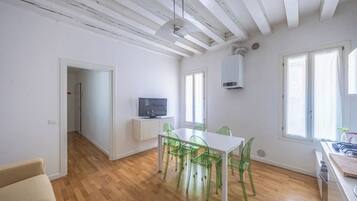 Un espacio habitable amplio y luminoso con techo de madera, una zona de comedor con mesa blanca y sillas verdes, un televisor sobre un mueble blanco y ventanas grandes que dejan entrar luz natural.