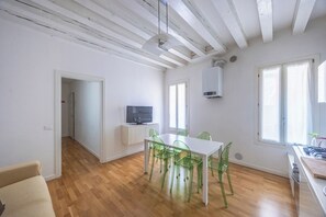Un espacio habitable amplio y luminoso con techo de madera, una zona de comedor con mesa blanca y sillas verdes, un televisor sobre un mueble blanco y ventanas grandes que dejan entrar luz natural.
