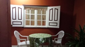 Un pequeño comedor con una mesa redonda cubierta con mantel verde, dos sillas blancas y una ventana con contraventanas blancas.