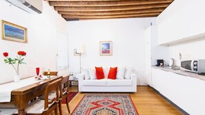 Un espace de vie compact comprenant un coin repas, un canapé blanc avec des coussins rouges, un tapis à motifs et un coin cuisine équipé d’un four à micro-ondes.