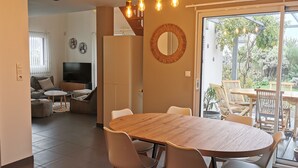 Una sala moderna con televisor, sofá y mesa de centro. Cuenta con una zona de comedor con una mesa de madera y sillas. La habitación tiene un amplio ventanal corredizo que da a un patio con mobiliario exterior.