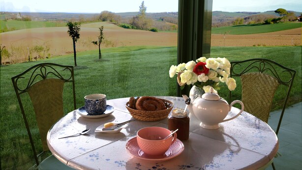 Een gedekte tafel met een theepot, kopjes, een kom en een mandje brood, met uitzicht op een prachtig landschap.