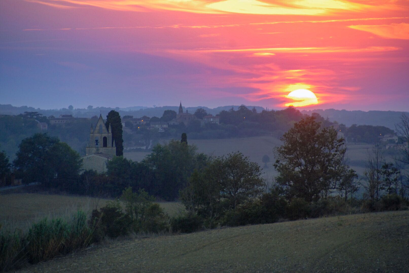 Een landelijk landschap met een kerk en velden bij zonsondergang.
