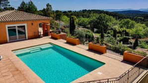 A swimming pool with a tiled deck, surrounded by planters and a fence, overlooking a lush landscape with trees and distant hills. - Villa Théia (Flayosc)