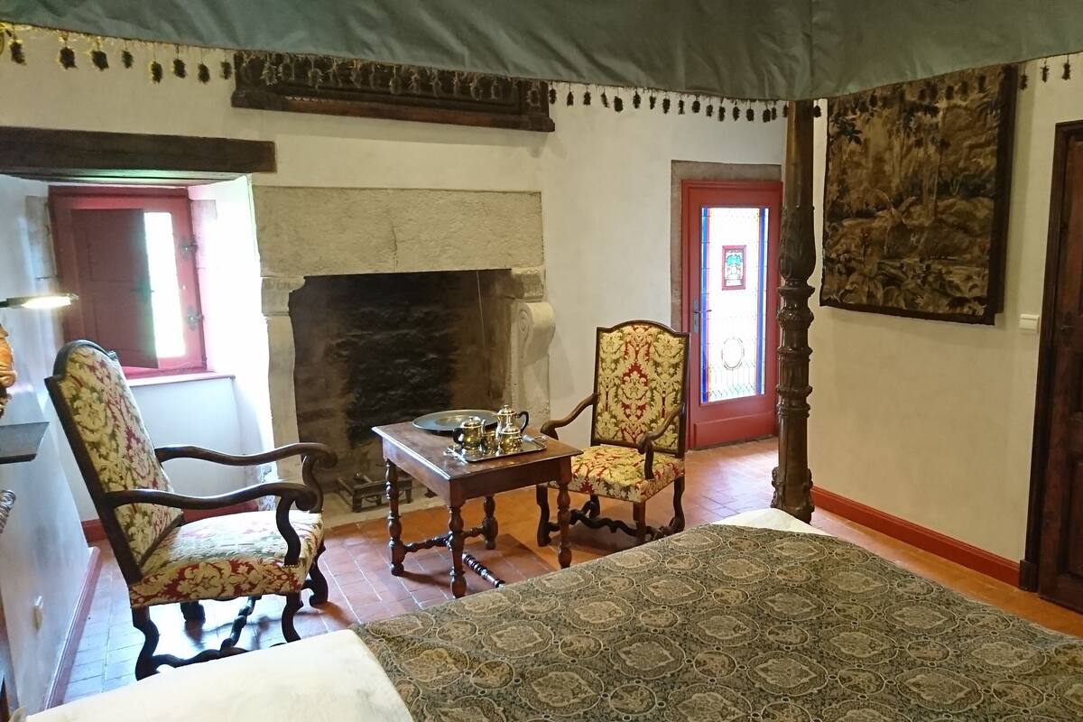 Ein traditionell eingerichteter Raum mit Kamin, einem Bett mit gemustertem Überwurf, zwei Polstersesseln, einem Holztisch mit Tee-Service und einem großen, gerahmten Gemälde an der Wand.