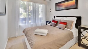 A neatly made bed with a beige and white duvet, a white towel, and several pillows in a modern bedroom with a large window, a ceiling fan, and a framed picture on the wall. - Ren Basera - Minutes from the base of Mt Buller (Sawmill Settlement)