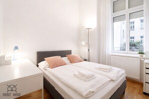 A neatly made bed with pillows, a bedside lamp, a small table, and a window with a view of buildings.