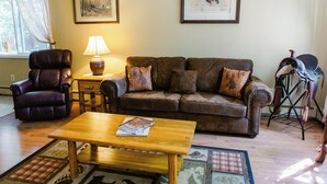 Un salón con un sillón de cuero, una mesa de centro de madera con libros, un sofá marrón con cojines, una lámpara y un cuadro en la pared.