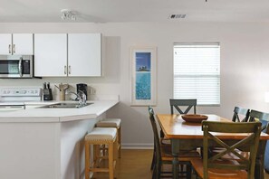 Una cocina con armarios blancos, un microondas y un lavamanos. Hay una mesa de comedor con sillas y una ventana con persianas.