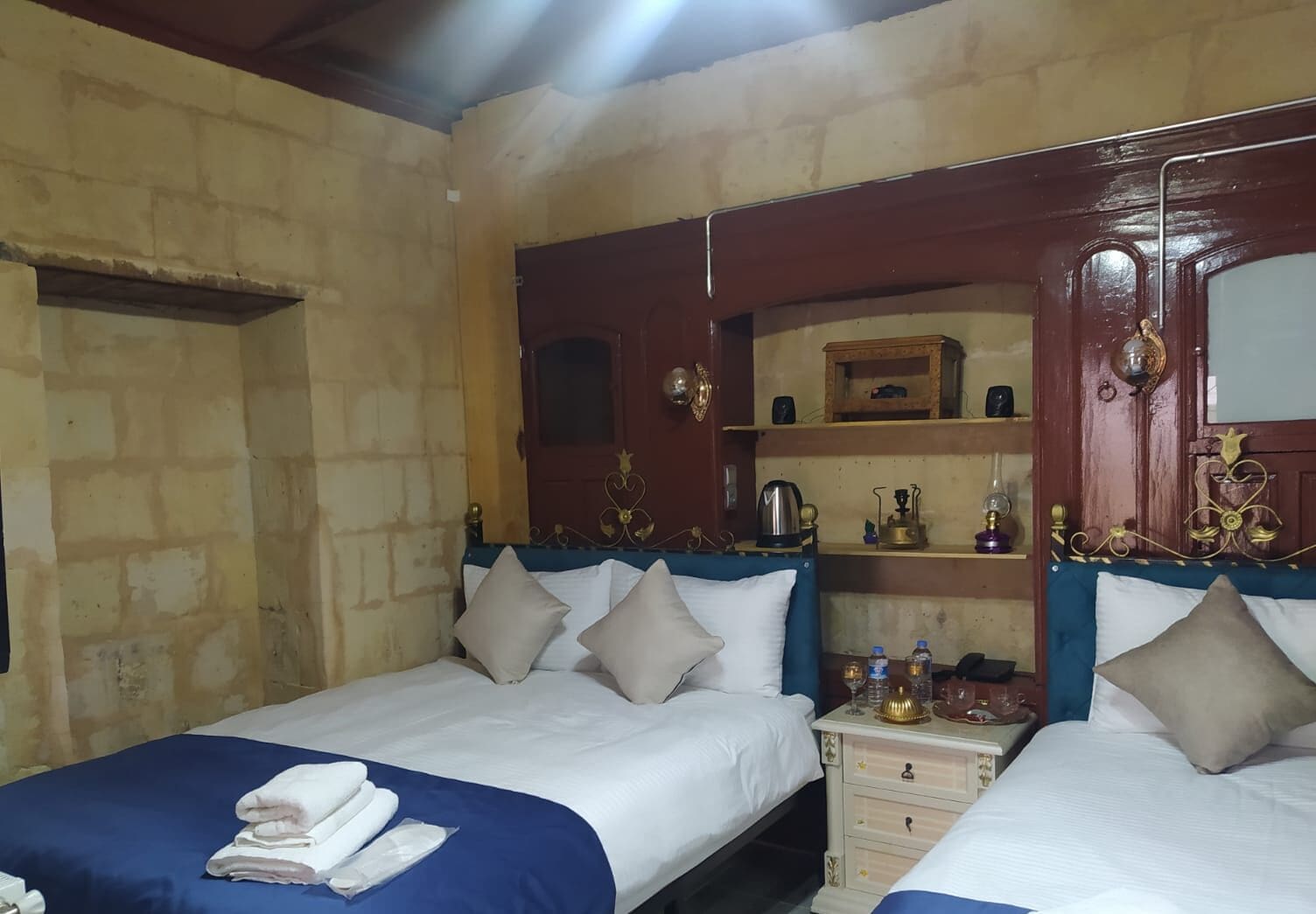 Habitación de hotel con dos camas, cada una con cubrecama azul y sábanas blancas. Cuenta con un cabecero de madera con detalles metálicos decorativos, un mueble de madera pequeño con espejo y una mesita de noche con lámpara y botella de agua.