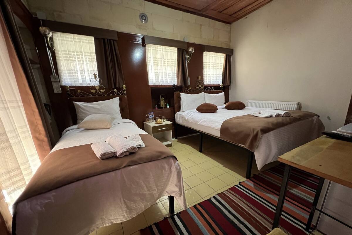 Ein Hotelzimmer mit zwei Einzelbetten, einem kleinen Tisch, einem Nachttisch und einem Fenster mit Vorhängen.