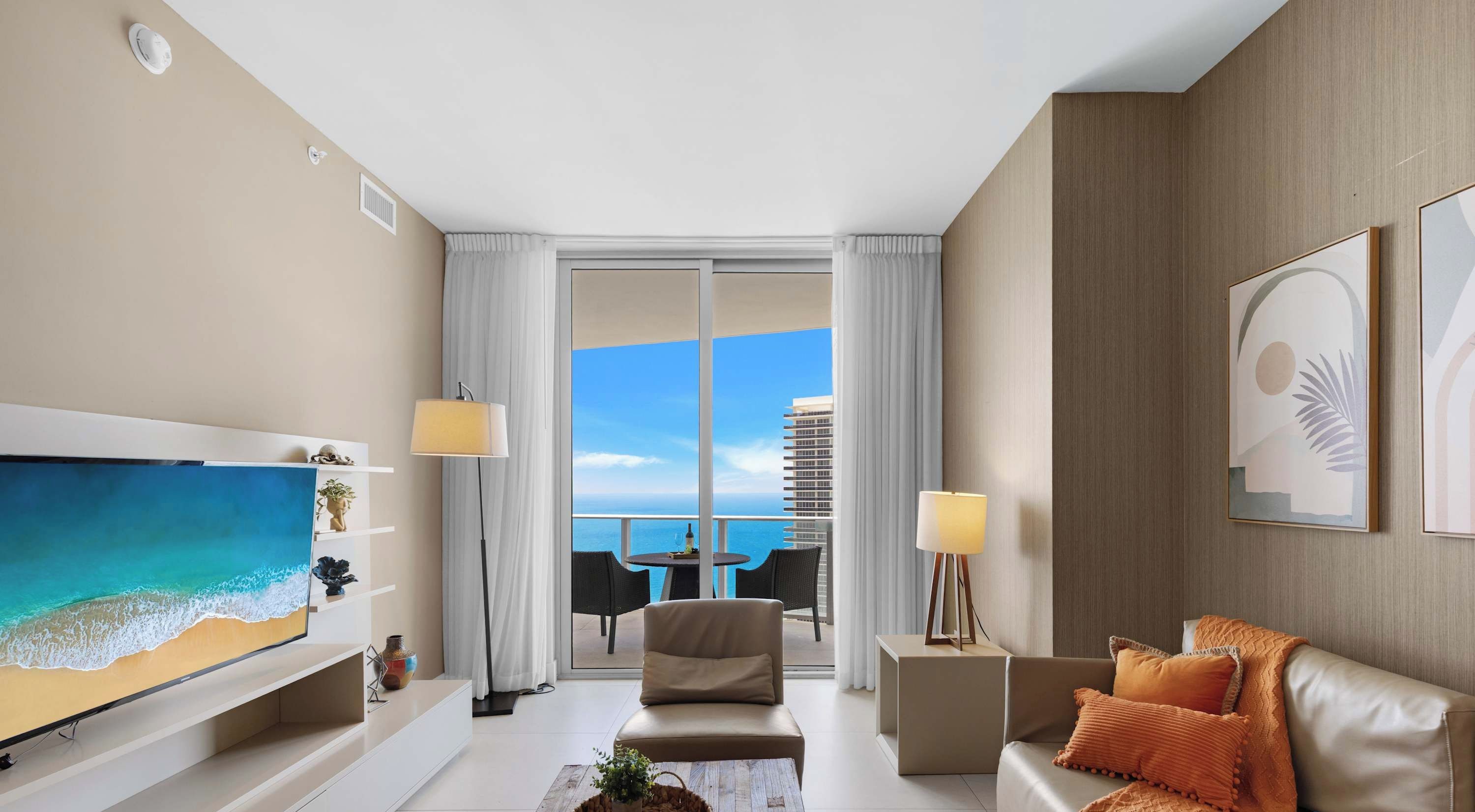 A modern hotel room with a large TV, a sofa, a coffee table, and a balcony with a view of the ocean.
