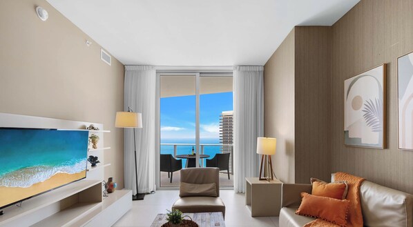 A modern hotel room with a large TV, a sofa, a coffee table, and a balcony with a view of the ocean.