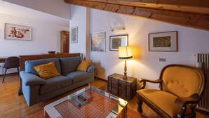 Un salon confortable avec un canapé, un fauteuil et une petite table. Des tableaux encadrés ornent le mur et une lampe est posée sur une table d’appoint.