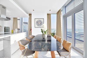 A modern kitchen with a white countertop, stainless steel appliances, and a dining area with a glass table and cushioned chairs. - Luxe 3BR with maids room in Bluewaters (Dubai)