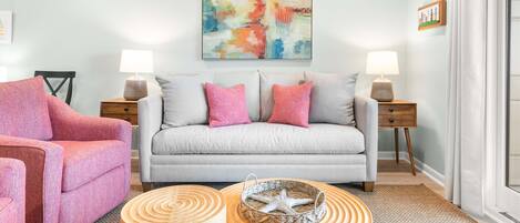 Een zithoek met een grijze bank, een roze fauteuil, twee ronde houten bijzettafeltjes en een kleurrijk abstract schilderij aan de muur.