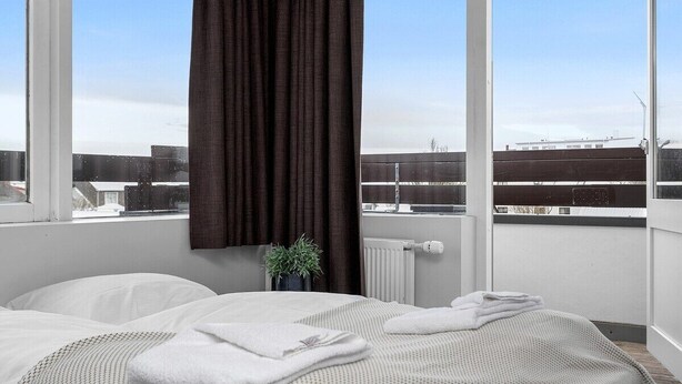 A bedroom with a large window, a bed with white linens, a dark curtain, a small plant, and a view of buildings. - Svala Downtown Studio (Reykjavík)
