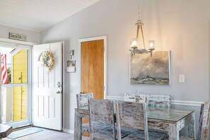 Un coin repas avec une table et des chaises en bois, un lustre, un tableau et une porte décorée d’une guirlande.