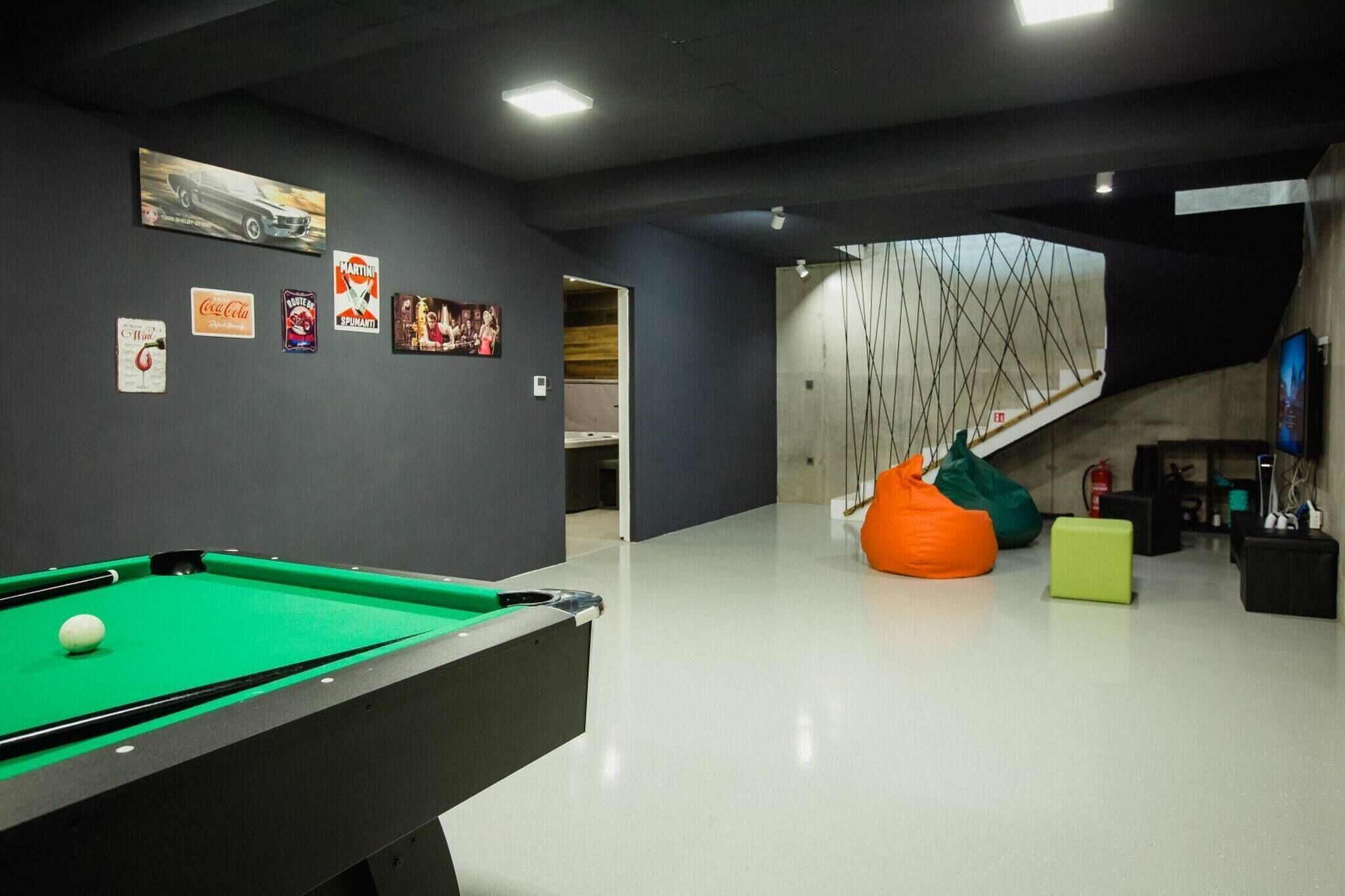 Mesa de bilhar com bola branca, mesa de sinuca e sacos de feijão em um ambiente moderno com escada e obras de arte nas paredes.
