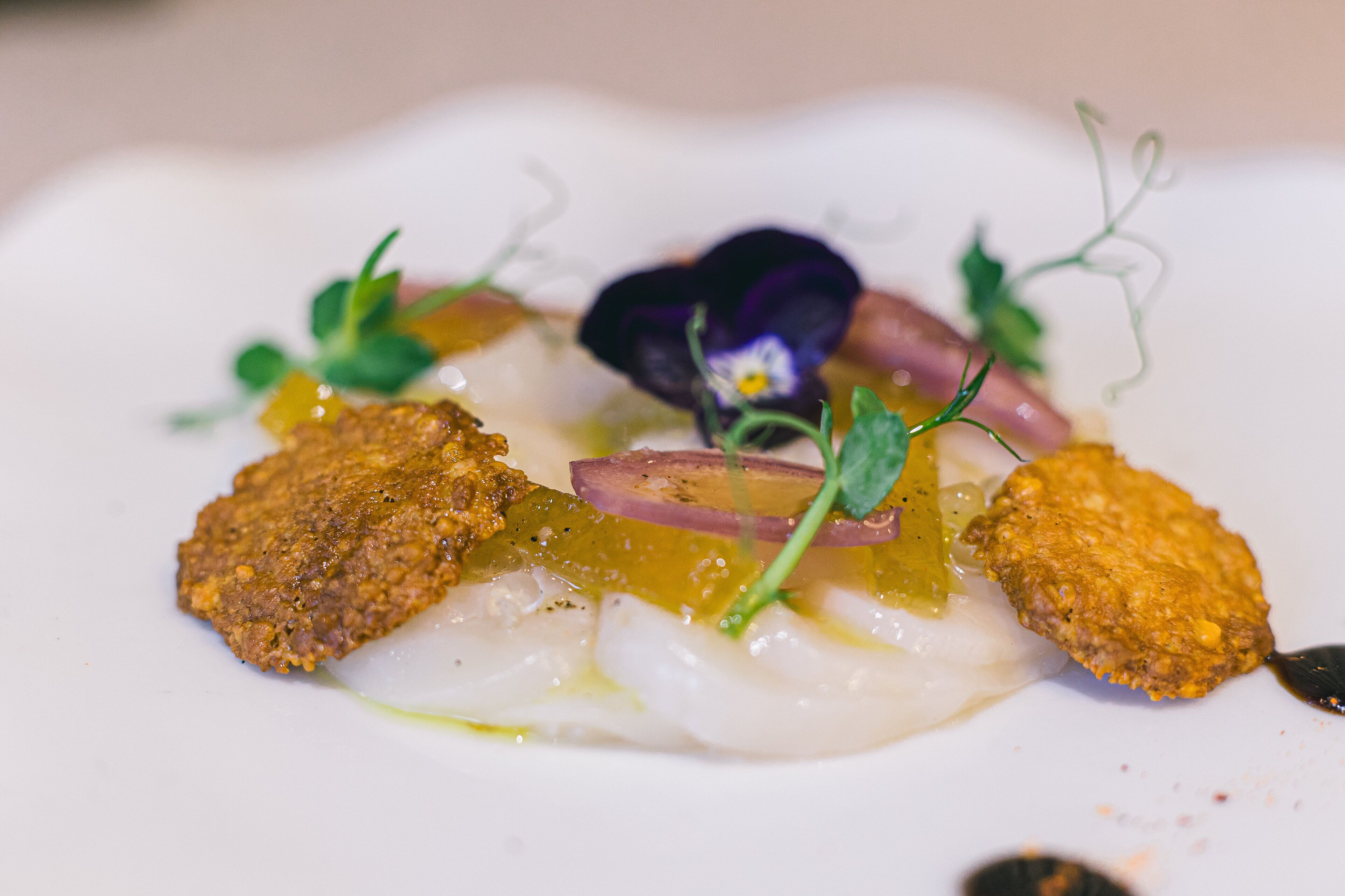 A dish with white fish, green herbs, and a purple flower.