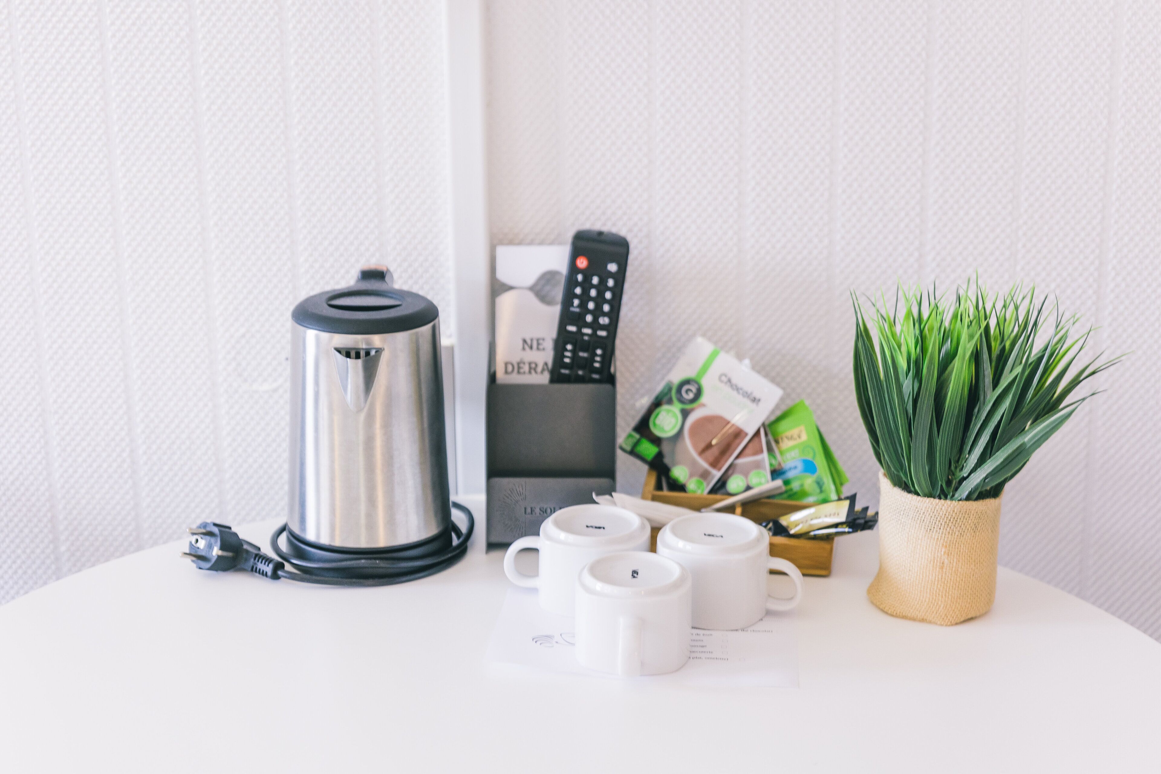 A white table with a stainless steel kettle, a remote control, a plant in a woven pot, and several white mugs.