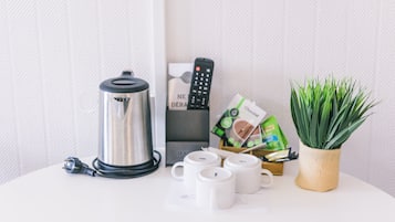 A white table with a stainless steel kettle, a remote control, a plant in a woven pot, and several white mugs.