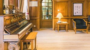 A piano, a wooden bench, a table with a lamp, and a chair in a room with wooden walls and a wooden floor. - Le Soleil D'or (Montigny-la-Resle)