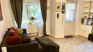 Un salon avec un canapé de couleur sombre, une petite table, un radiateur, une porte et une étagère ornée d’objets décoratifs.