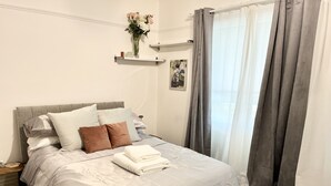 Un lit bien fait, avec des oreillers, une table de chevet, une commode, un vase de fleurs, un cadre photo et une fenêtre avec des rideaux.