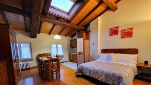 Quarto com cama de madeira, área de refeições com mesa e cadeiras, cozinha ao fundo e um claraboia acima da cama.