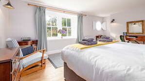 A hotel room with a large bed, a desk with a chair, a wooden dresser, a window with curtains, a bedside table with a lamp, and a mirror on the wall.