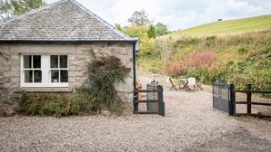 A stone cottage with a gravel driveway, a wooden gate, and a chair outside.