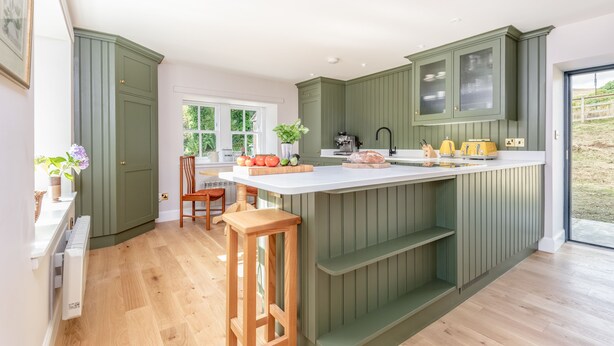 A kitchen with green cabinets, a white countertop, and a wooden stool.