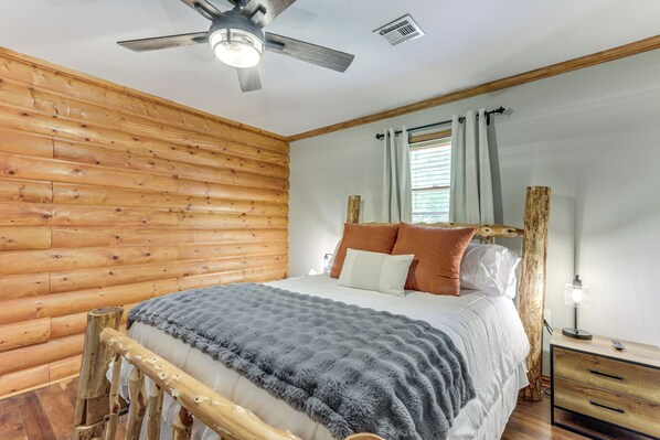 Un dormitorio de cabaña de troncos con cabecera de madera, una cama con manta gris, una mesita de noche con lámpara y una ventana con cortinas.