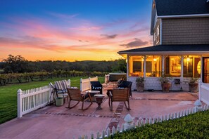 A patio with outdoor furniture, a hot tub, and a view of a vineyard at sunset.