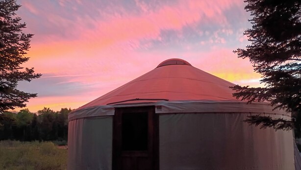 A yurt with an open door, surrounded by rocks and trees, under a sunset sky. - Redarrow rustic yurt  in the Adirondacks,  Gore Mt. 15 mins, jug water supplied (Minerva)